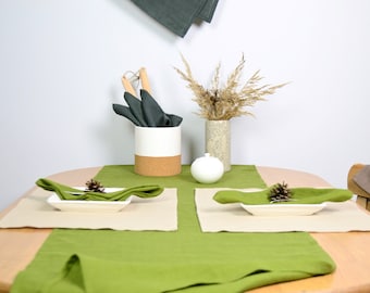 May include: A dining table setting featuring a green table runner, two beige placemats, and two white rectangular plates topped with green napkins and small pinecones. A white and cork utensil holder, a white bowl, and a vase with dried grass.