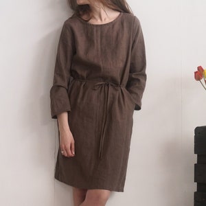 May include: A dark brown knee-length dress with long sleeves, a round neckline, and a matching fabric tie belt at the waist. The garment features a simple, straight-cut silhouette with visible side pockets, crafted from a textured fabric.