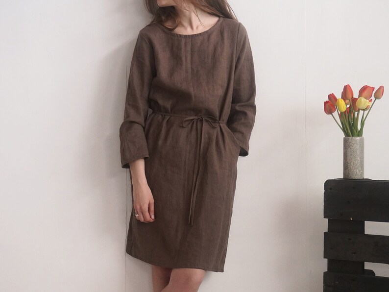 May include: A dark brown knee-length dress with long sleeves, a round neckline, and a matching fabric tie belt at the waist. The garment features a simple, straight-cut silhouette with visible side pockets, crafted from a textured fabric.