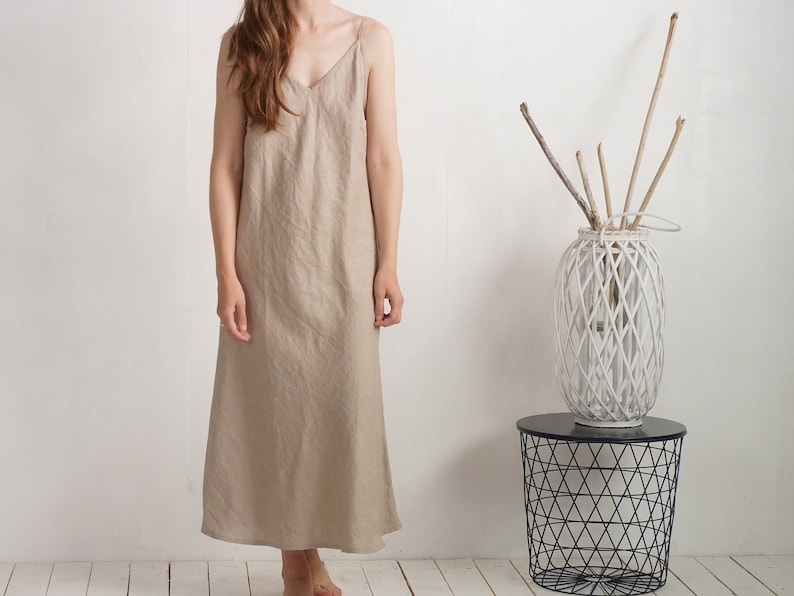 Op de afbeelding: Een enkellange mouwloze jurk in beige met een V-hals en dunne spaghettibandjes. Het kledingstuk heeft een ontspannen, recht model dat tot op de enkels valt, gemaakt van een gestructureerde stof met een zichtbaar natuurlijk weefpatroon.