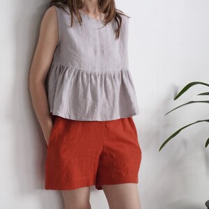 Puede incluir: Una blusa sin mangas de color gris claro con cuello redondo y cintura con volante fruncido, combinada con pantalones cortos de color naranja quemado. La blusa tiene un corte holgado y los pantalones cortos tienen un corte recto con bolsillos.