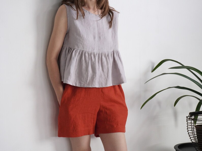 Puede incluir: Una blusa sin mangas de color gris claro con cuello redondo y cintura con volante fruncido, combinada con pantalones cortos de color naranja quemado. La blusa tiene un corte holgado y los pantalones cortos tienen un corte recto con bolsillos.