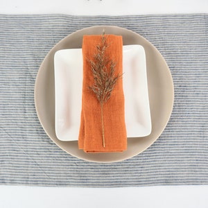 May include: A white square plate with a beige round plate on top. An orange linen napkin is folded and placed on the plates with a dried branch of grass on top. The plates are on a blue and white striped linen placemat.