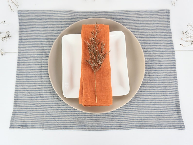 May include: A white square plate with a beige round plate on top. An orange linen napkin is folded and placed on the plates with a dried branch of grass on top. The plates are on a blue and white striped linen placemat.