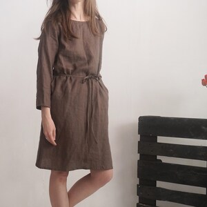 May include: A knee-length brown dress with long sleeves, a round neckline, and a thin matching tie belt at the waist. The garment features a simple, straight-cut silhouette with a visible seam running vertically down the centre of the bodice.