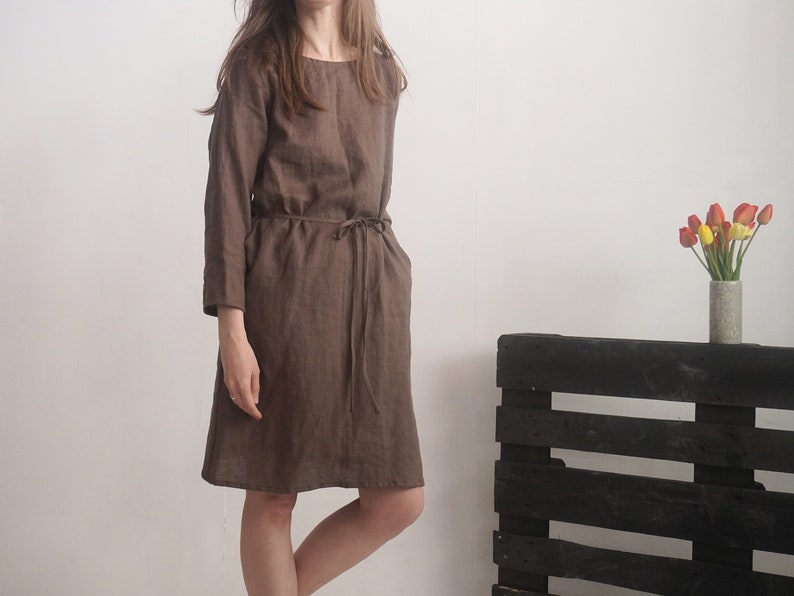 May include: A knee-length brown dress with long sleeves, a round neckline, and a thin matching tie belt at the waist. The garment features a simple, straight-cut silhouette with a visible seam running vertically down the centre of the bodice.