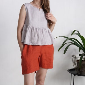 Puede incluir: Una blusa sin mangas de color lavanda claro con volante en la cintura y cuello redondo, combinada con pantalones cortos de color naranja quemado. La blusa presenta una cintura fruncida que crea una silueta acampanada y bolsillos laterales.