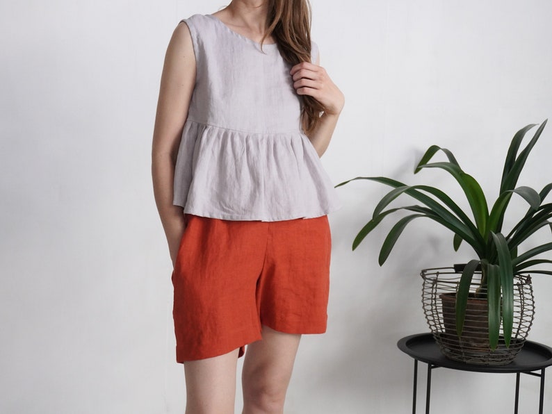 Puede incluir: Una blusa sin mangas de color lavanda claro con volante en la cintura y cuello redondo, combinada con pantalones cortos de color naranja quemado. La blusa presenta una cintura fruncida que crea una silueta acampanada y bolsillos laterales.