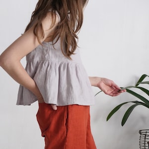 Puede incluir: Una blusa sin mangas de color lavanda claro con cintura fruncida y cuello redondo, combinada con pantalones de color naranja quemado liso con bolsillos laterales. La blusa est&aacute; confeccionada en un tejido texturizado y llega a la cadera.