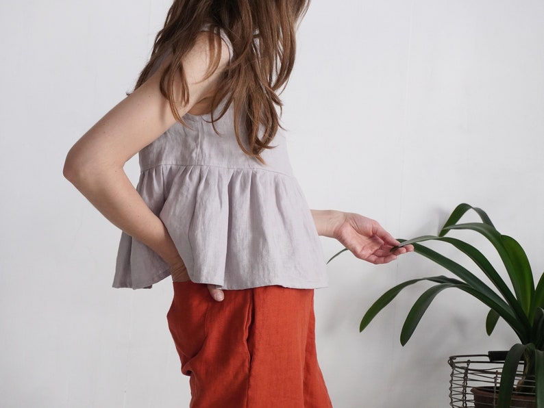 Puede incluir: Una blusa sin mangas de color lavanda claro con cintura fruncida y cuello redondo, combinada con pantalones de color naranja quemado liso con bolsillos laterales. La blusa est&aacute; confeccionada en un tejido texturizado y llega a la cadera.