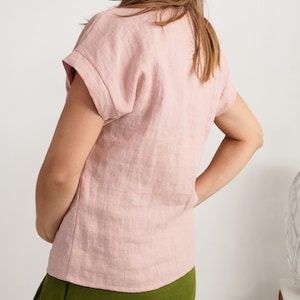 May include: A light pink short-sleeved top with a textured fabric finish and cuffed sleeves. The garment features a simple round neckline and a straight hemline, providing a clean and straightforward look for everyday wear.