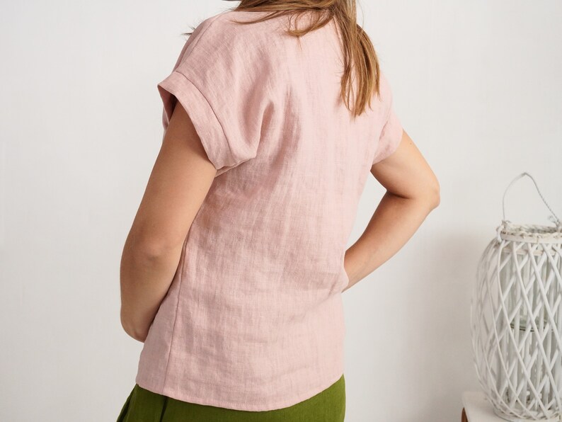 May include: A light pink short-sleeved top with a textured fabric finish and cuffed sleeves. The garment features a simple round neckline and a straight hemline, providing a clean and straightforward look for everyday wear.