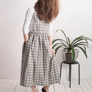 May include: A long pinafore dress featuring a grey and beige checkered pattern, a gathered elastic waist, and wide shoulder straps with button closures. The dress is worn over a plain white long-sleeved shirt and includes side pockets.