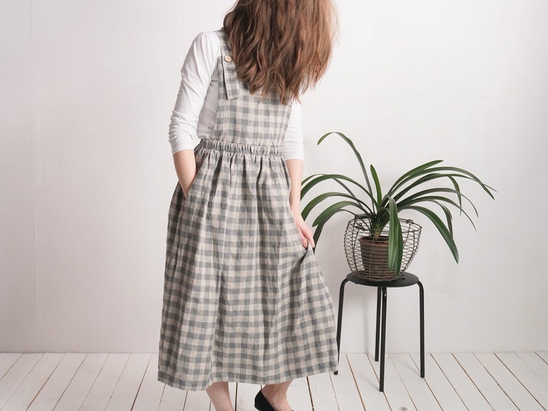 May include: A long pinafore dress featuring a grey and beige checkered pattern, a gathered elastic waist, and wide shoulder straps with button closures. The dress is worn over a plain white long-sleeved shirt and includes side pockets.