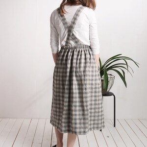 May include: A midi-length pinafore skirt featuring a grey and beige checkered pattern. The garment has a gathered elasticized waistband and wide shoulder straps that cross over the back, worn over a plain white long-sleeved shirt.