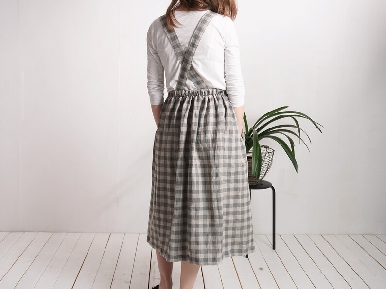May include: A midi-length pinafore skirt featuring a grey and beige checkered pattern. The garment has a gathered elasticized waistband and wide shoulder straps that cross over the back, worn over a plain white long-sleeved shirt.