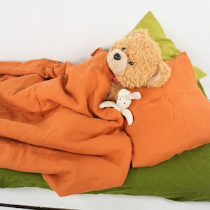 May include: A plush brown teddy bear with a white bunny toy lies in a bed with a rust-colored linen duvet and a green linen pillow.