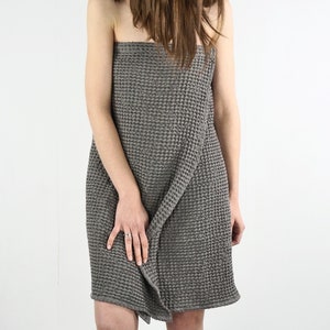 May include: A gray waffle weave towel wrapped around a person's body. The towel is a light gray color and has a waffle texture. The person is standing in front of a white background.