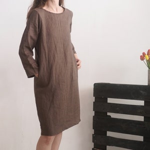 May include: A knee-length brown dress with long sleeves, a round neckline, and side pockets. The garment features a simple, straight-cut silhouette and is crafted from a textured fabric, providing a neutral and understated appearance for daily wear.