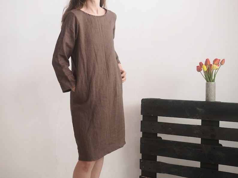 May include: A knee-length brown dress with long sleeves, a round neckline, and side pockets. The garment features a simple, straight-cut silhouette and is crafted from a textured fabric, providing a neutral and understated appearance for daily wear.