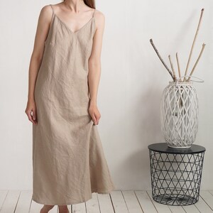 Op de afbeelding: Een beige enkellange slipdress met een V-hals en dunne spaghettibandjes. Het kledingstuk heeft een los, recht model dat tot op de enkels valt en is gemaakt van een effen stof met een zichtbare, natuurlijke weefstructuur.