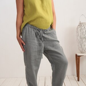 May include: A person wearing a casual outfit. The outfit consists of a sleeveless, olive green linen top and grey linen trousers with a drawstring waist. The trousers are cuffed at the ankles. The person is wearing white trainers.
