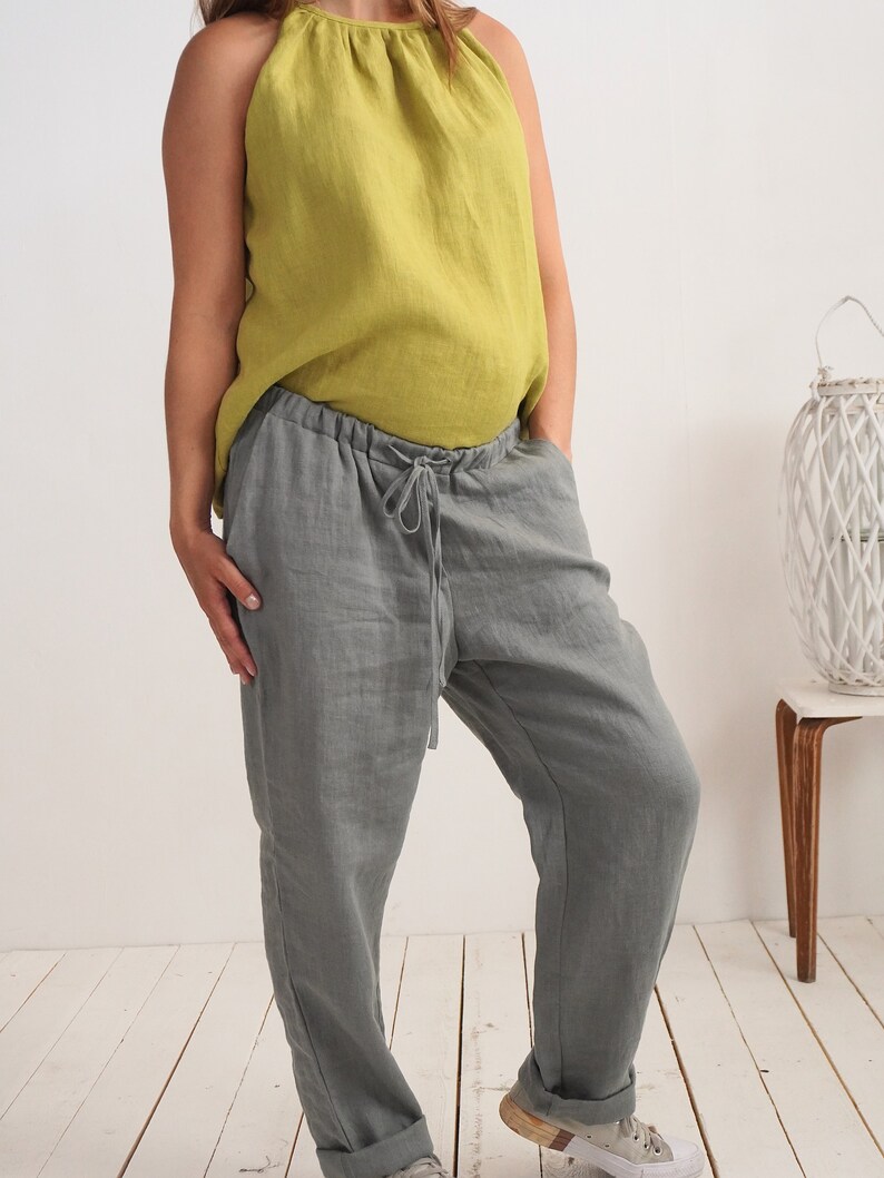 May include: A person wearing a casual outfit. The outfit consists of a sleeveless, olive green linen top and grey linen trousers with a drawstring waist. The trousers are cuffed at the ankles. The person is wearing white trainers.