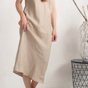 Op de afbeelding: Een mouwloze midi-jurk in een effen lichtbeige kleur. Het kledingstuk heeft een eenvoudig, losvallend silhouet met een ronde halslijn en een rechte zoom, gemaakt van een gestructureerde stof met zichtbare weefdetails.