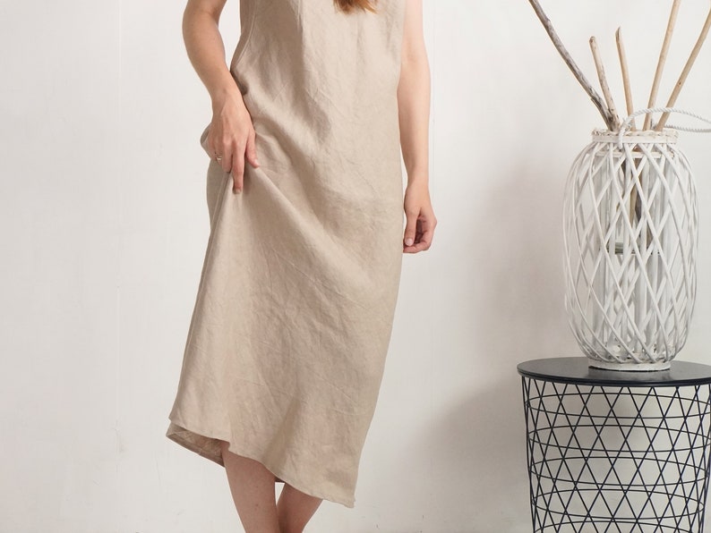 Op de afbeelding: Een mouwloze midi-jurk in een effen lichtbeige kleur. Het kledingstuk heeft een eenvoudig, losvallend silhouet met een ronde halslijn en een rechte zoom, gemaakt van een gestructureerde stof met zichtbare weefdetails.