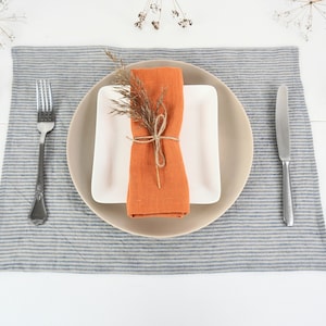 May include: A place setting with a white plate, a beige plate, and an orange linen napkin tied with twine. The napkin is decorated with a sprig of dried flowers. The place setting is on a blue and white striped linen placemat.