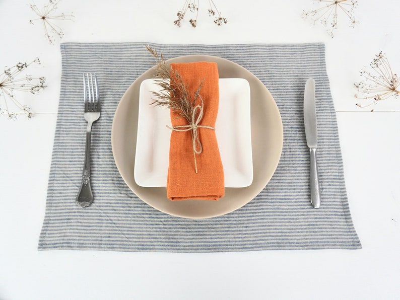 May include: A place setting with a white plate, a beige plate, and an orange linen napkin tied with twine. The napkin is decorated with a sprig of dried flowers. The place setting is on a blue and white striped linen placemat.