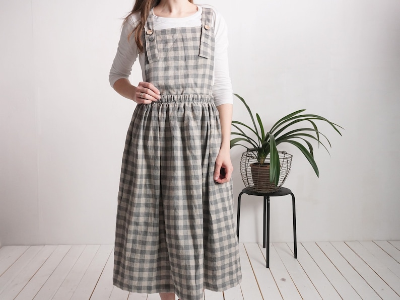 May include: A pinafore dress featuring a grey and beige gingham check pattern, a gathered elasticized waist, and wide shoulder straps secured with light-colored wooden buttons, worn over a plain white long-sleeved shirt.