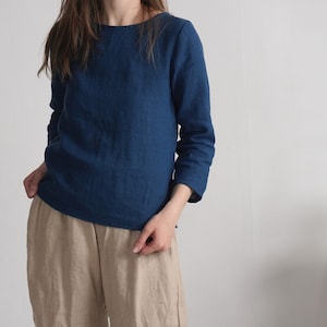 May include: A long-sleeved, deep blue top with a round neckline, paired with loose-fitting beige trousers. The top features a straight hemline and three-quarter length sleeves, crafted from a textured fabric that appears to be linen.