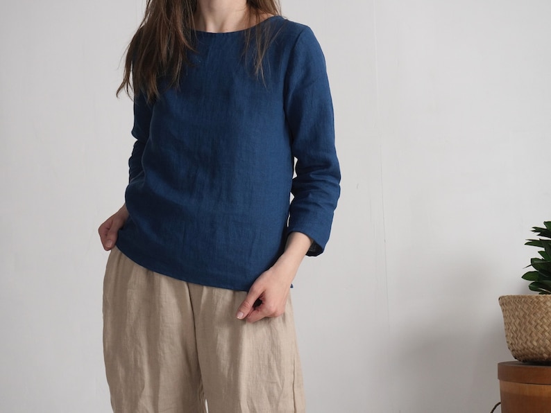 May include: A long-sleeved, deep blue top with a round neckline, paired with loose-fitting beige trousers. The top features a straight hemline and three-quarter length sleeves, crafted from a textured fabric that appears to be linen.