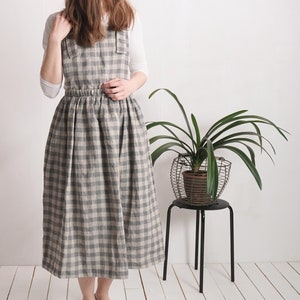 May include: A pinafore dress featuring a grey and beige checkered pattern with a square neckline, wide shoulder straps, and a gathered elasticized waistline that flows into a full-length skirt, worn over a plain white long-sleeved shirt.