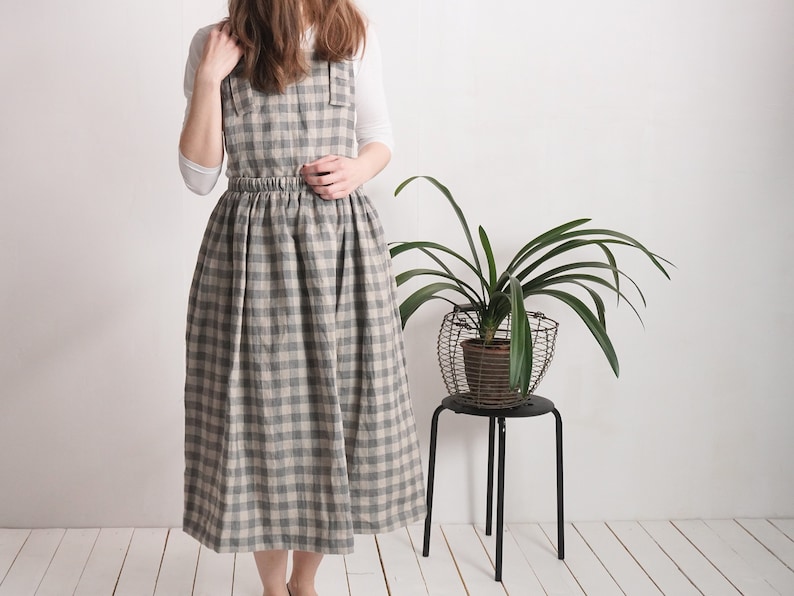 May include: A pinafore dress featuring a grey and beige checkered pattern with a square neckline, wide shoulder straps, and a gathered elasticized waistline that flows into a full-length skirt, worn over a plain white long-sleeved shirt.