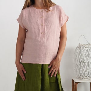 May include: A light pink short-sleeved linen blouse featuring a round neckline and a front placket with four light-colored buttons. The blouse has cuffed sleeves and a straight hemline, paired with a solid olive green skirt.