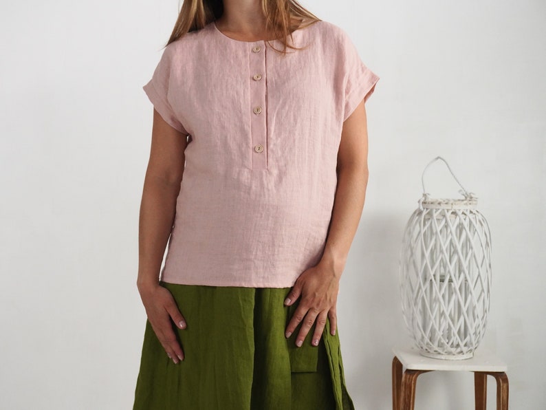May include: A light pink short-sleeved linen blouse featuring a round neckline and a front placket with four light-colored buttons. The blouse has cuffed sleeves and a straight hemline, paired with a solid olive green skirt.