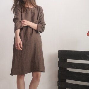 May include: A knee-length brown dress with a round neckline and three-quarter length sleeves. The garment features a loose, straight-cut silhouette and is crafted from a textured fabric, falling just above the knees with a simple hemline.