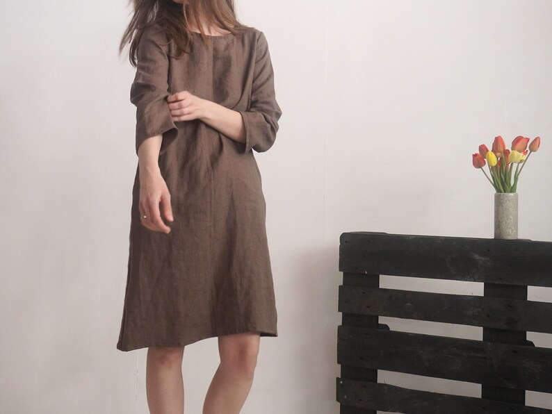 May include: A knee-length brown dress with a round neckline and three-quarter length sleeves. The garment features a loose, straight-cut silhouette and is crafted from a textured fabric, falling just above the knees with a simple hemline.