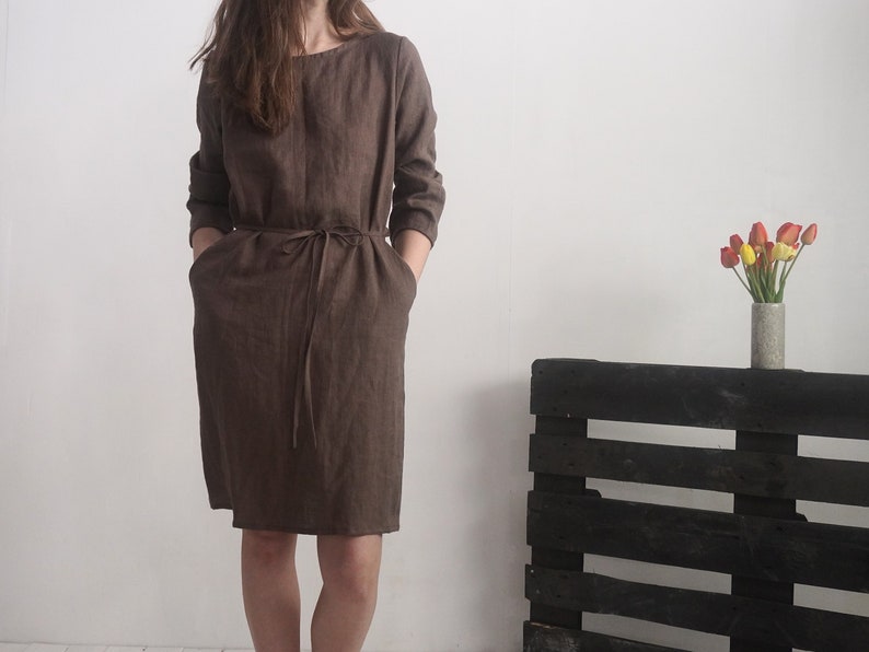 May include: A knee-length, dark brown dress with long sleeves, a round neckline, and a matching fabric tie belt at the waist. The garment features two side pockets and is constructed from a textured fabric with visible weave details.