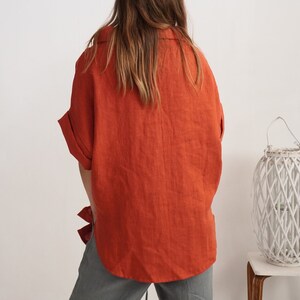 Puede incluir: Una camisa abotonada de color naranja quemado con cuello y mangas cortas con dobladillo, combinada con pantalones grises. La camisa presenta un corte holgado y un dobladillo curvo, vista desde atr&aacute;s, con un farol blanco al lado.
