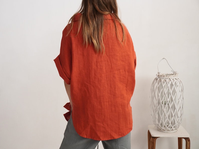 Puede incluir: Una camisa abotonada de color naranja quemado con cuello y mangas cortas con dobladillo, combinada con pantalones grises. La camisa presenta un corte holgado y un dobladillo curvo, vista desde atr&aacute;s, con un farol blanco al lado.