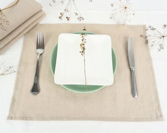 Puede incluir: Un mantel individual de lino beige con un plato cuadrado blanco y un plato verde debajo. Un tenedor y un cuchillo de plata se colocan a cada lado del plato. Una ramita de flores secas se coloca en el plato.