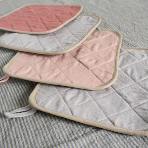 May include: Four quilted pot holders stacked on top of each other. The top two are pink and the bottom two are grey. The pot holders have a beige trim.