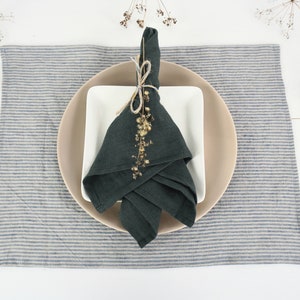 May include: A dark green linen napkin folded and tied with twine, decorated with dried flowers, on a white square plate, on a beige round plate, on a blue and white striped linen placemat.