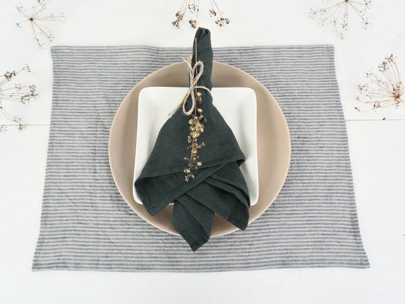 May include: A dark green linen napkin folded and tied with twine, decorated with dried flowers, on a white square plate, on a beige round plate, on a blue and white striped linen placemat.
