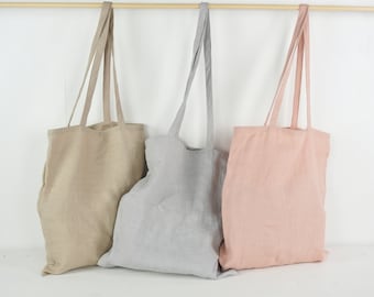 May include: Three tote bags hanging from a wooden rod. The bags are made of a plain fabric in light beige, light gray, and soft pink. Each bag features two long shoulder straps and a simple rectangular shape with a visible woven texture.