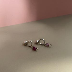 以下が含まれることがあります： 小さな金属製の球体が円形のリングに取り付けられたシルバーのスタッドピアス一対。それぞれのフープから、ファセットカットが施された濃い紫色のビーズが一つずつぶら下がっています。ピアスは磨き上げられた仕上げで、シンプルな構造です。
