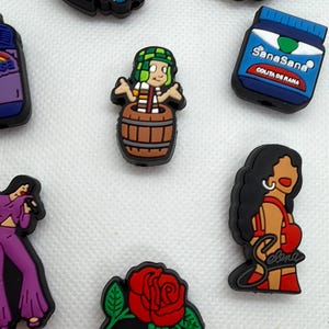 May include: A collection of colorful shoe charms featuring various designs. Included are a Hello Kitty figure, a Frida Kahlo portrait, a cartoon character in a barrel, a blue box with text, a woman in a red dress, and a rose with text.